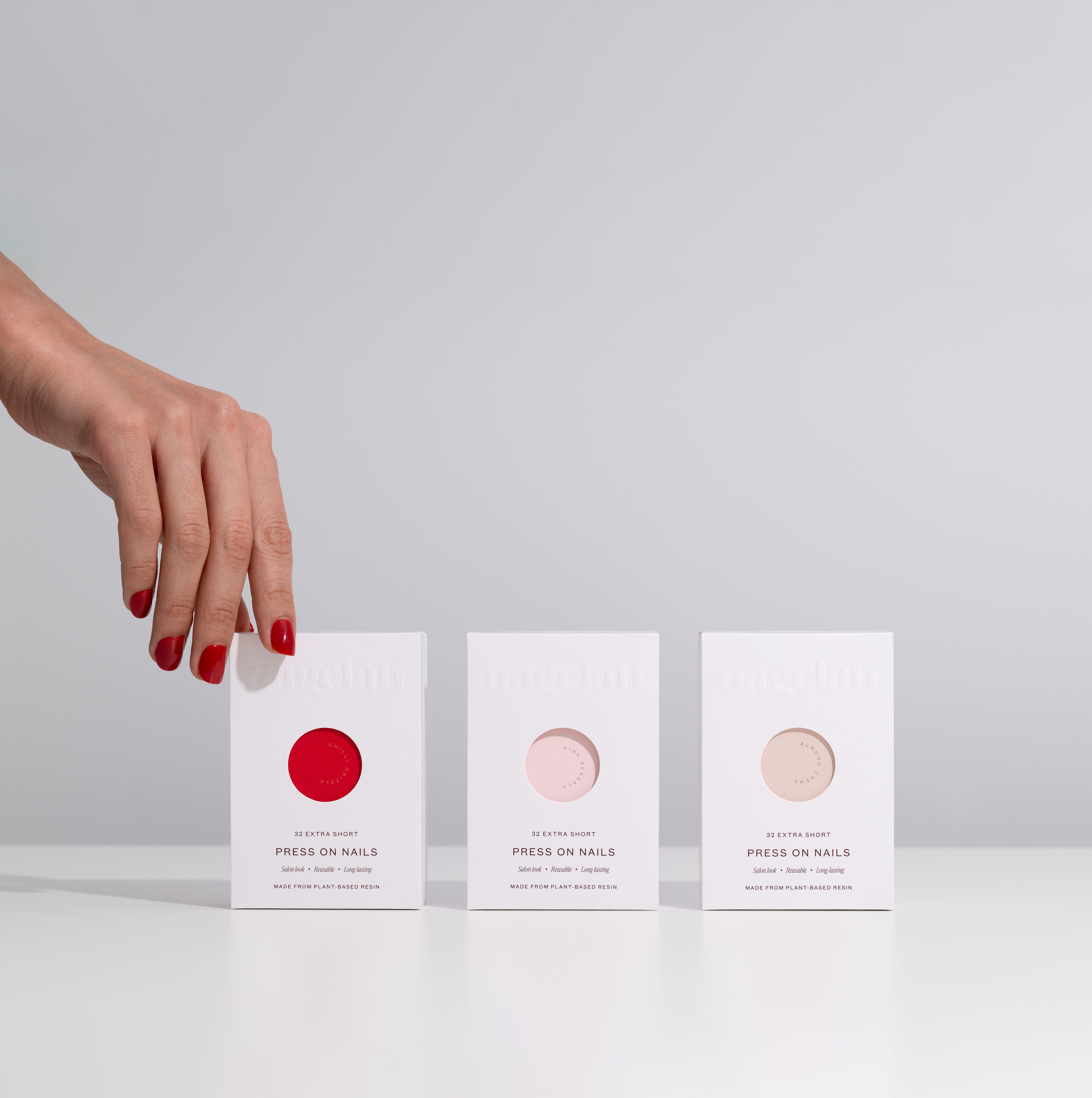 Three boxes of press-on nails held by a hand with red nail polish on a light gray background